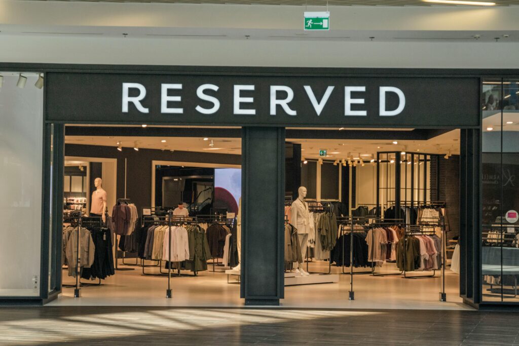 The entrance of a modern retail clothing store named 'Reserved,' displaying mannequins and neatly arranged clothing racks, representing fashion retail and shopping experience